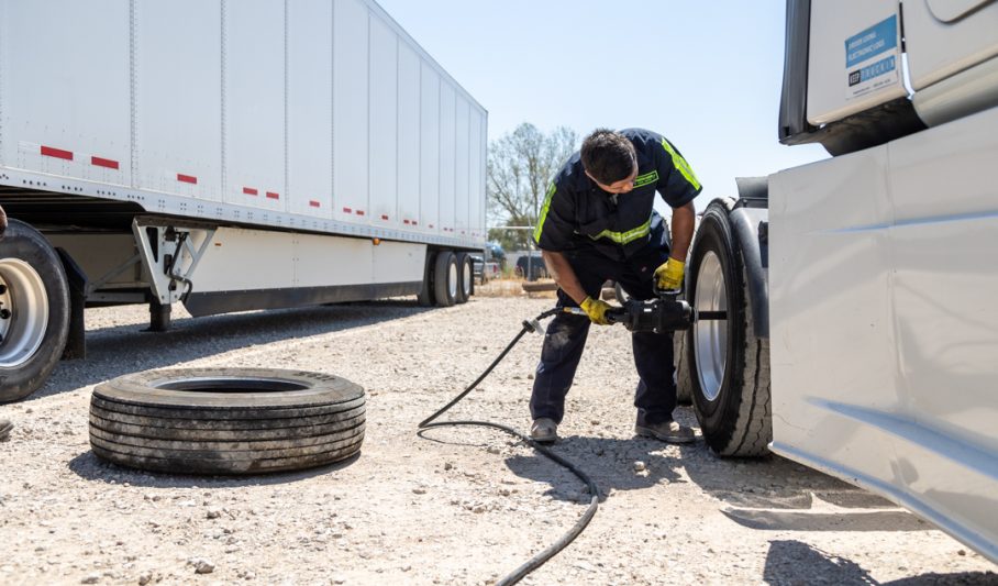 A technician using an air impact wrench to change a semi truck tire outdoors, a worn spare tire lying on the gravel beside him and a trailer parked behind