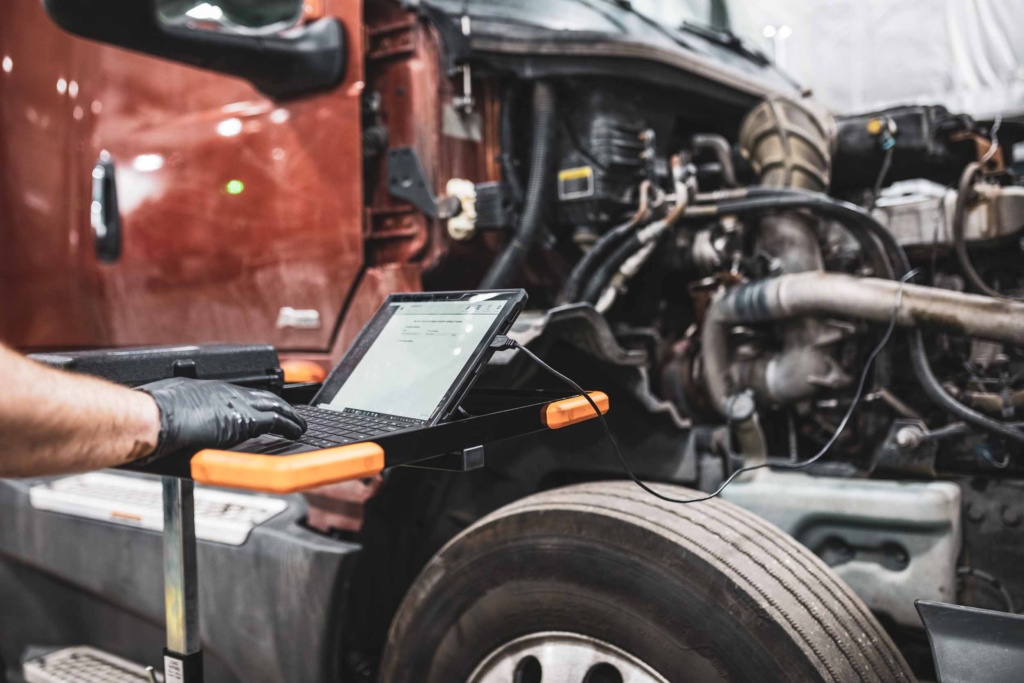 A technician running diagnostics on a rugged laptop plugged into a semi truck's exposed engine