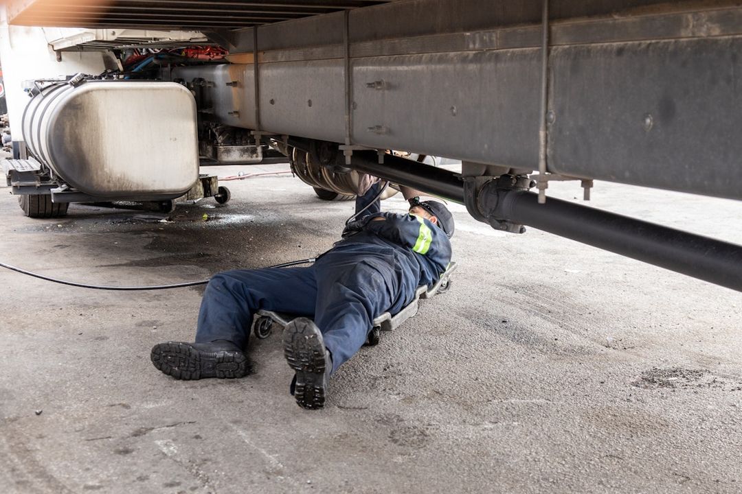 A technician on a creeper working underneath a truck in a yard, repairing its underside and driveline