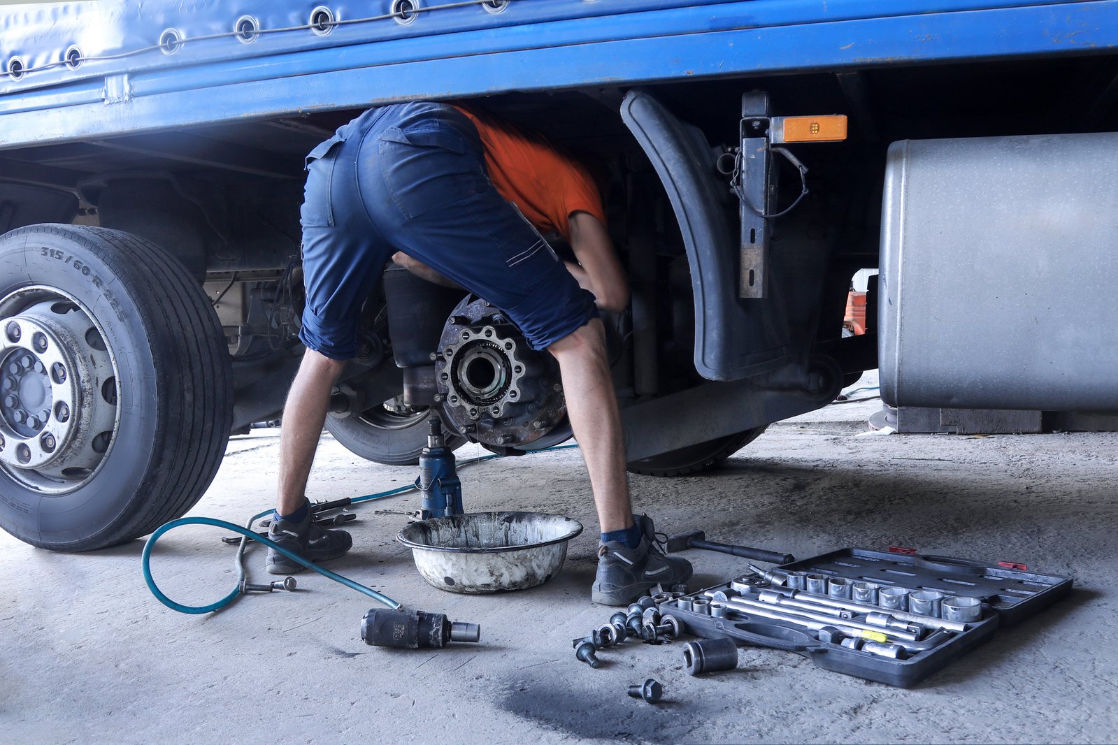 A mechanic working on a truck's exposed brake hub with the wheel removed, sockets, impact wrench and tools laid out on the ground
